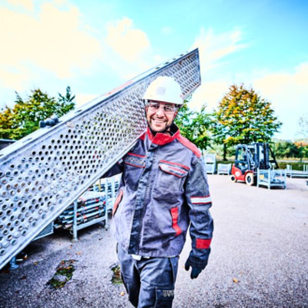 Lächelnder KAEFER-Mitarbeiter mit Schutzhelm trägt ein langes Gitterrost-Element für den Gerüstbau auf der Schulter