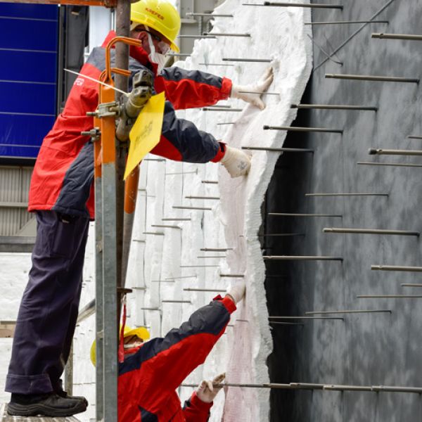 Ein Arbeiter mit Atemschutzmaske und Schutzbrille bringt Dämmmatten an einer Wand an, die durch zahlreiche horizontal herausstehende Metallstifte fixiert werden