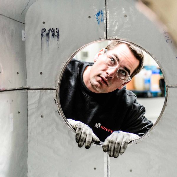 KAEFER Mitarbeiter mit Schutzbrille blickt durch eine runde Öffnung in einer hellen Industriewand